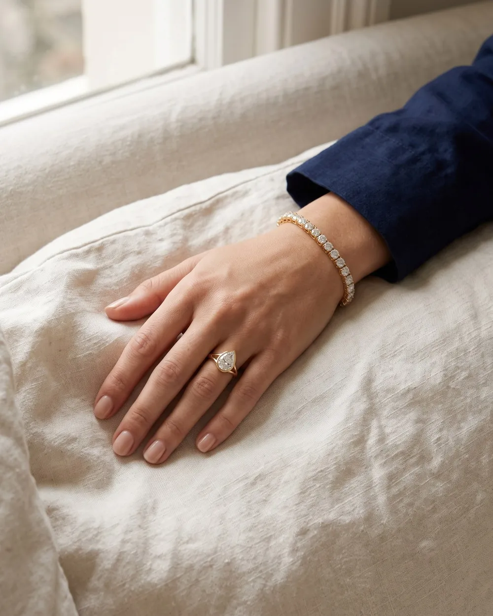 A hand resting on linen, wearing an Aaryam diamond tennis bracelet and a pear bezel ring